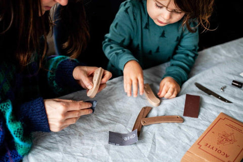 Photo d'une maman et son enfant qui font une activité manuelle autour d'une table avec une nappe blanche. Ils Poncent tous les 2 leurs manches en bois d'un couteau à assembler