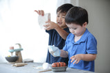 2 garçons de 3 ans qui sont entrain de jouer avec une dinette en bois représentant un set à pâtisserie. Le 1er garçon a un gant de cuisine et semble sortir un moule du four et le 2eme soulève un sachet de farine