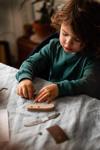 Photo d'un jeune enfant qui est assit au bord d'une table, il porte un pull over vert et il est concentré sur une activité manuelle