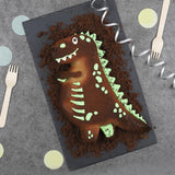 Photo pris de haut d'une table de fête d'anniversaire avec posé sur une planche un gâteau au chocolat en forme de dinosaure. Il est décoré avec des pointes de couleurs verte et blanche