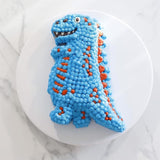 Photo d'un gâteau dinosaure recouvert d'un glacage bleu rt rouge. Le gateau est posé sur une assiette blanche, elle même posée sur un plan de travail en marbre