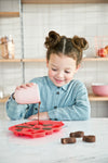 Photo d'une petite fille dans une cuisine. La fille transverse du chocolat dans un moule de couleur rouge. Devant le moule est posé 3 petits gâteaux au chcolat