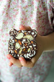 Gros plan sur la main d'une petite fille tenant dans sa main un gâteau au chocolat recouvert de plusieurs fruits sec. La petite fille porte un chemisier au motif liberty rose