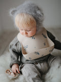 Jeune enfant blond avec une cagoule grise sur la tête qui porte des vêtements en laine avec un bavoir beige avec des motifs fleurs lilas desu. La photo a un style retro