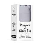Emballage en carton blanc de son set keepies et transport de voyage de couleur gris. L'emballage laisse entrevoir le dessus de l'etui avec des yeux et oreilles d'ours. Les inscriptions sur l'emballage sont en anglais, il y a é pictogrammes qui précise que le produit convient a un usage alimentaire et l'autre que le produit résiste a une température entre -40° et 230°. L'ensemble est disposé sur un fond blanc.