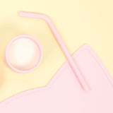 Gros plan sur une paille en silicone de couleur rose. La paille est posée sur une table de couleur jaune pâle. A coté de la paille il y a un gobelet de la même couleur avec du lait a l'intérieur. La photo est prise par le dessus