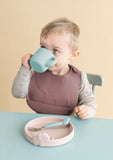 Peek phto d'un garçon qui boit dans une tasse de couleur verte, il a posé devnt lui une assiette en silicone rose avec une fourchette à l'intérieur. Le petit garçin a un bavoir en silicone bordeaux autour du cout