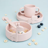 Peek photo d'une table bleue clair avec posé dessus de la vaisselle pour enfant en silicone, il y a un bol, une assiette et une tasse de la meme couleur rose