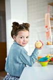 Photo d'une jeune fille qui regarde l'objectif avec dans une de ces main un gâteaux avec déssiné dessus la tête d'un ours. La fille a le coude posé sur une table avec à coté 3 petits moules de couleurs empilés les un aux autres