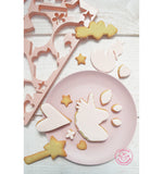Biscuit sur le theme des licorne posé sur une assiette rose pastel et sur la table. il y a sur le coté de la photo une plaque de découpoir rose pastel