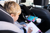 Jeune enfant assis sur un siege auto dans une voiture, il a une tablette devant lui avec de la nourriture de posé. il tient dans sa main un stylo et il est entrain de dessiner