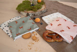  Photo d’une table en bois ou est posé dessus des pochettes à snack en tissus contenant des goûter dont un bretzel. Les pochettes sont carrés et de couleur differentes