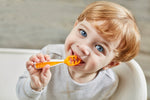Photo d'un petit garçon, blond vénisien aux yeux bleu et souriant. Il porte a sa bouche une cuillère de couleur orange. Il est sourient et est habillé d'un tee shirt aux couleurs claires
