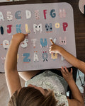 Photo prise en hauteur d'une table avec posée dessus un set de table coloré avec des illustration de lettre d'alphabet rigolos. Devant le set est assis 2 enfants que l'ont apperçoit et qui montent des lettre sur le set de table