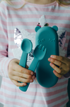 Gros plan sur les main d'un enfant tenant une fourchette et une cuillère en silicone de couleur bleu, dans l'autre main il tient un étuis de transport pour les couvert de la même couleur et en forme de tête d'ours endormis
