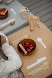 Phto d'un set de table posé sur une table avec des illustration de vaisselle dessus. Sur l'emplacement de l'assiette il y a un bol avec des fruits à l'intérieur, sur l'emplacement des couverts il y a une fourchette et une cuillère à soupe. Sur le coté haut gauche du set de table il y a une girafe