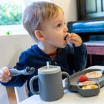 Jeune garçon assit à table entrain de manger des fruits. Il tient dans sa man une petite fourchette avec des myrtilles dessus. Devant lui sont posés de la vaisselle de couleur grise