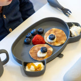 Photo d'une assiette pour enfant en forme de tête d'ours. Dans l'assiette il y a une repas, les aliments sont disposés de façon à former un visage. il y a des pancakes, des fruits rouges et du yaourt