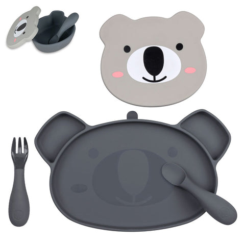 Set de repas pour enfant composé de plusieurs éléments. Le tout est posé sur un fond blanc. Il y a en avant plan une assiette compartimentée en silicone de couleur grise, l’assiette à une forme de koala. Une cuillère et une fourchette de la même couleur sont posés dans et à côté de l’assiette. Au dessus de l’assiette, le couvercle d’un bol de couleur gris et de forme de tête de koala avec des illustrations coloré. Sur le côté gauche un bol de la même couleur et de forme de tête de Koala.