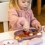Photo d'une jeune fille blonde assise à table. Elle mange dans de la vaisselle posée devant elle. La jeune fille à un pul en maille rose. La vaisselle est rose pale