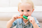 Bébé blond qui regarde l'objectif et porte a sa bouche 2 brosses à dents en silicone de couleur verte et orange
