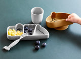 Photo d'une table de couleur bleu indigo avec posé dessus 2 assiette en silicone pour enfant, 1 est en forme de crocodile avec 3 compartiment et contenant des fruits, l'autre est un bol de couleur jaune moutarde. La main d'un enfant essaie dedecoler le bol de la table. Il y a également un gobelet ainsi qu'une fourchette de la même couleur et texture que l'assiette crocodile