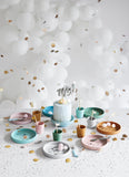 A Phto d'une table de fête blanche avec des ballon et des confettis couleur or. Sur la table est posées en rond de la vaisselle en silicone de couleurs differentes