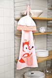 Tablier et toque de cuisine pour enfant posés sur une étagère de cuisine, avec de la vaisselle dedans. L'ensemble est de couleur rose pale avec une illustration de renard qui fait la cuisine dessus