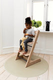 Photo d'une jeune fille qui mange un gateau, elle est asisse sur une chaise haute en bois et tient un doudou entre ses jambes. La chaise est posée sur un tapis de sol rond de couleur beige