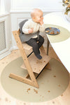 Bébé assi sur une chaise haute en bois devant une assiette grise, il a la main devant sa bouche et beaucoup de miettes est éparpillés sur le sol et un tapis rond de coueur beige