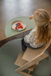 Phot prise ne hauteur d'une jeune fille assise a table sur une chaise haute. Il y a une assiette devant elle avecdes fruits rouges à l'intérieur. L'assiette est posée sur un tapis en silicone en forme de demi cercle de couleur vert olive. Au sol il y a un tapis de la même couleur