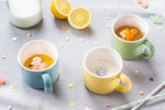 Table avec une nape grise et des cotillons de couleurs dessus. Sur la table il y a 3 tasses en porcelaine avec des animaux a l'intérieur. Une est bleu avec un flamand rose dedans, l'autre jaune avec un éléphant et la dernière verte avec un écureuil