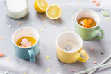 Table avec une nape grise et des cotillons de couleurs dessus. Sur la table il y a 3 tasses en porcelaine avec des animaux a l'intérieur. Une est bleu avec un flamand rose dedans, l'autre jaune avec un éléphant et la dernière verte avec un écureuil