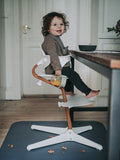 Jeune enfant assit sur une chaise haute, devant une table à manger. La photo est prise sur le coté de la chaise, l'enfant est habillé d'un pull en maille et il tient dans sa main une tasse d'apprentissage avec du jus d'orange à l'intérieur