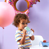 Photo d'un jeune garçon au cheveux bouclés qui boit dans une gourde. Derrière lui il y a un mur violet avec 2 gros balon, un rond et l'autre en forme de donuts grignoté