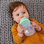 bebe au regard dans le vide entrain de boire dans une gourde au couvercle vert d'eau. Le bébé est allongés sur un tapis gris au grosse maille. Le bébé porte un tee shirt orange