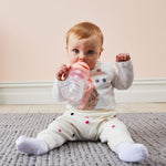 Photo d'un bébé assit par terre sur un tapis, qui est entrain de boire dans une gourde au couvercle rose. Le bébé a les cheveux roux et il porte des affaires au ton clair