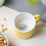 Tasse en porcelaine de couleur jaune avec a l'intérieur un elephant gris en porcelaine. L'intérieur de la tasse est blanche et il y a du lait dedans. La tasse est posée sur une table avec des cacahuètes a coté