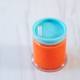 Photo d'un gobelet pour enfant contenant un liquide orange. La tasse à une base transparente, elle est refermée par un couvercle de couleur vert, le couvercle à une paille transparente 