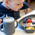 Jeune garçon assi a table devant une assiette en silicone de couleur grise avec à l'intérieur des pancakes décoré avec des fruits rouges. A coté de son assiette il y a une tasse de la même couleur