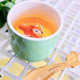 Tasse en porcelaine de couleur verte avec a l'intérieur un écureuil orange en porcelaine. L'intérieur de la tasse est blanche et il y a du jus de pomme dedans. La tasse est posée  sur une table avec devant la tasse une cuillère en bois.