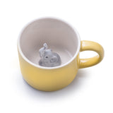 Tasse en porcelaine de couleur jaune avec a l'intérieur un elephant gris en porcelaine. L'intérieur de la tasse est blanche. La tasse est posée  sur un fond blanc