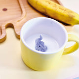 Tasse en porcelaine de couleur jaune avec a l'intérieur un elephant gris en porcelaine. L'intérieur de la tasse est blanche. La tasse est posée sur une table, a l'intérieur il y a du lait et on voit sur le fond floutté de la photo une assiette en bambou en forme d'elephant avec une banane posée dessus