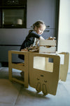 Photo d'une jeune fille brune assit sur un bureau en bois clair, elle est en pleine activité avec une caisse dejouet posée devant elle sur le bureau