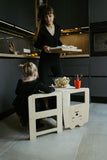 Photo prise dans une cuisine de couleur grise avce une jeune fille blonde assise a un petit bureau d'enfant en bois avec posé dessus des crayons de couleur, derrière il y a une femme adulte qui porte un plateau 