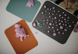 Photo prise en hauteur d'une table blanche avec posée dessus 3 set de table de couleurs differentes avec de la pate à modeler rose dessus