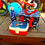 Boite à goûter, gourde et sac isotherme pour enfant posé sur un banc en bois avec en arrière plan un vélo pour enfant. L'ensemble est de couleur bleu nuit avec des illustrations de fusée et d'espace.