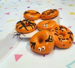 Donuts d'halloween aux couleurs orange avec des decors aux couleurs noir. Il y a une araignée en plastiqye sur le coté gauche de l'assiette ou sont posés les donuts