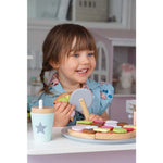 Jeune fille qui sourie, ele est entrain de jouer avec une pizza à decouper en bois posée devant elle. Elle porte une chemise bleu avec de smotifs de poissons de couleurs rose