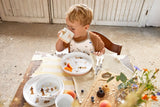 Photo d'un jeune garçon à table entrain de manger. Il y a sur la table de la jolie vaisselle pour enfant avec des fruits et gâteaux